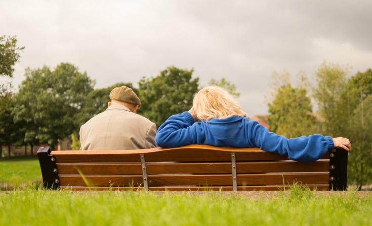 An elderly man and a woman sit on a park bench with their backs to the camera.